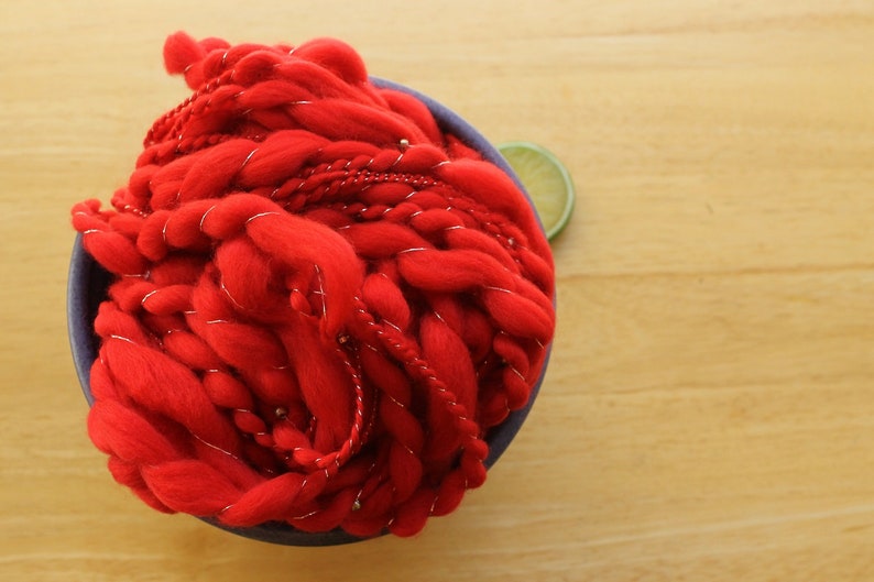 A hank of handspun, solid red, thick and thin yarn. The super bulky yarn is plied with silver thread and glass beads. It is nestled in a blue, ceramic yarn bowl on a light wood background with a lime slice.