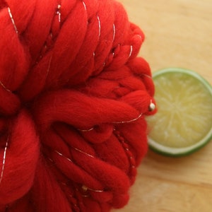 An extreme close up of a skein of handspun, solid red, thick and thin yarn. The super bulky yarn is plied with silver thread and glass beads. It is resting on a light wood background with a lime slice.