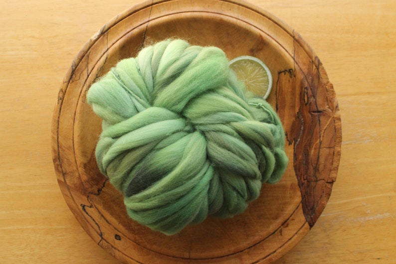 A skein of thick and thin, super bulky, handspun yarn. The yarn is hand dyed in dusty greens. It is resting on a handmade, wooden plate on a light wood background with a lime slice.