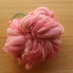 A skein of super bulky, hand dyed, handspun, thick and thin yarn. The yarn is bubblegum pink with wool curls. It is laying on a light wood background with a lime slice.