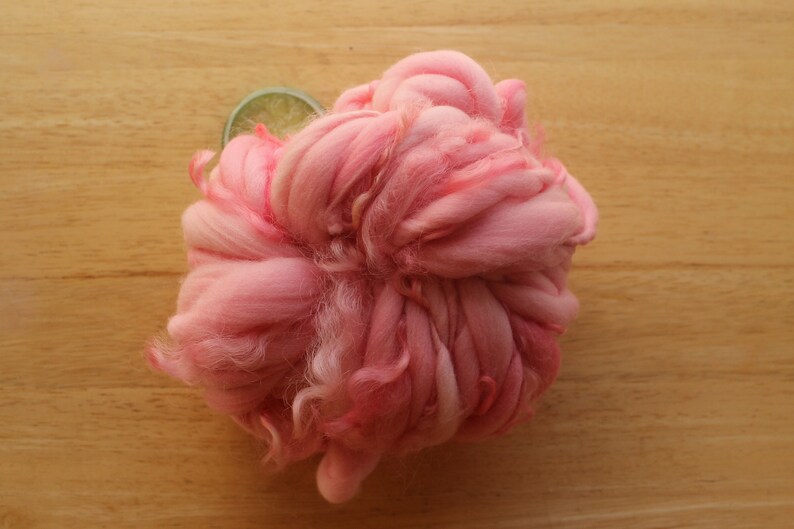 A skein of super bulky, hand dyed, handspun, thick and thin yarn. The yarn is bubblegum pink with wool curls. It is laying on a light wood background with a lime slice.