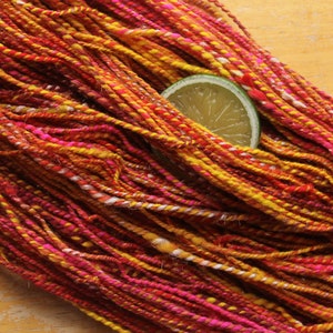 A hank of handspun, worsted weight, 2 ply yarn in red, hot pink, yellow, and white.  The yarn is resting on a light wood background with a lime slice.