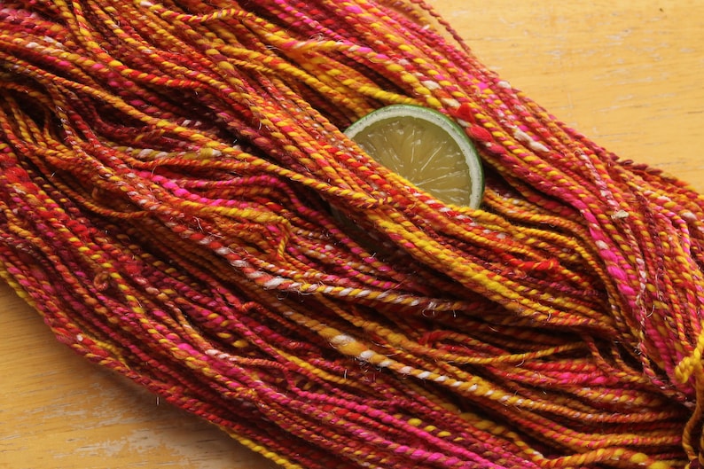 A hank of handspun, worsted weight, 2 ply yarn in red, hot pink, yellow, and white.  The yarn is resting on a light wood background with a lime slice.