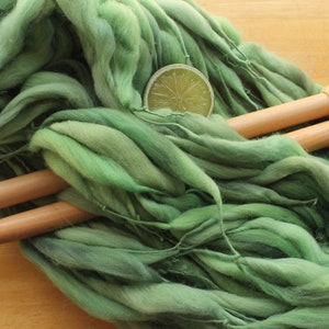 A hank of thick and thin, super bulky, handspun yarn. The yarn is hand dyed in dusty greens. It is resting on a light wood background with a pair of large, wooden knitting needles and a lime slice.