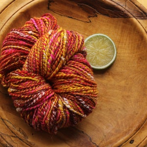 A skein of handspun, worsted weight, 2 ply yarn in red, hot pink, yellow, and white.  The yarn is resting on a handmade, wooden plate on a light wood background with a lime slice.