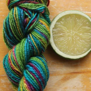 May include: A skein of colorful yarn in shades of green, teal, purple, and yellow sits next to a halved lime. The yarn is tightly wound, showcasing the vibrant color variations. The lime's interior is a pale yellow, with a green rind.