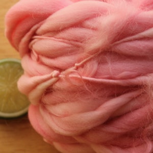 An extreme close up of a skein of super bulky, hand dyed, handspun, thick and thin yarn. The yarn is bubblegum pink with wool curls. It is laying on a light wood background with a lime slice.