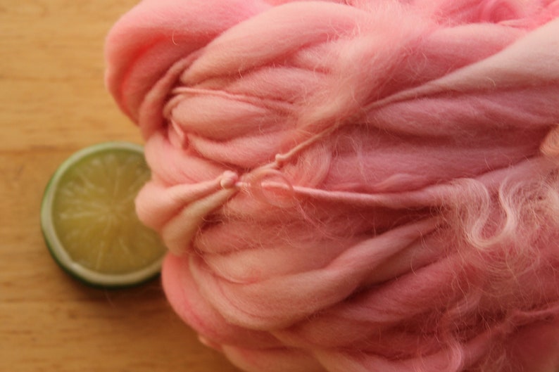 An extreme close up of a skein of super bulky, hand dyed, handspun, thick and thin yarn. The yarn is bubblegum pink with wool curls. It is laying on a light wood background with a lime slice.