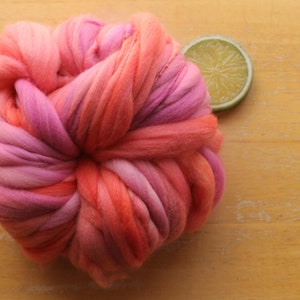 A skein of handspun, super bulky, thick and thin yarn. The yarn is hand dyed in self striping lilac and coral. The yarn is resting on a light wood background with a lime slice.