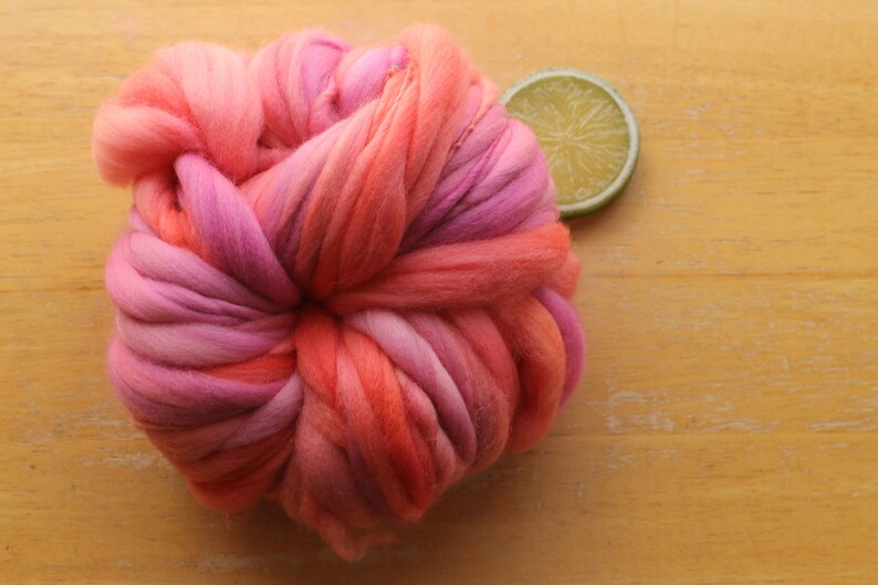 A skein of handspun, super bulky, thick and thin yarn. The yarn is hand dyed in self striping lilac and coral. The yarn is resting on a light wood background with a lime slice.