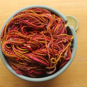 A hank of handspun, worsted weight, 2 ply yarn in red, hot pink, yellow, and white.  The yarn is nestled in a pale blue, ceramic yarn bowl on a light wood background with a lime slice.