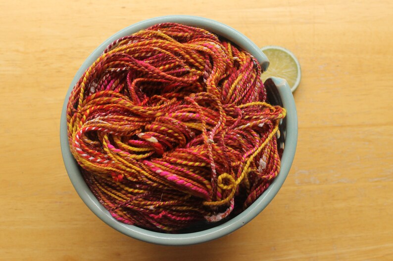 A hank of handspun, worsted weight, 2 ply yarn in red, hot pink, yellow, and white.  The yarn is nestled in a pale blue, ceramic yarn bowl on a light wood background with a lime slice.