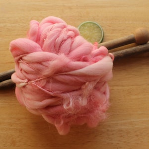 A skein of super bulky, hand dyed, handspun, thick and thin yarn. The yarn is bubblegum pink with wool curls. It is on a light wood background with a pair of large, wooden knitting needles and a lime slice.
