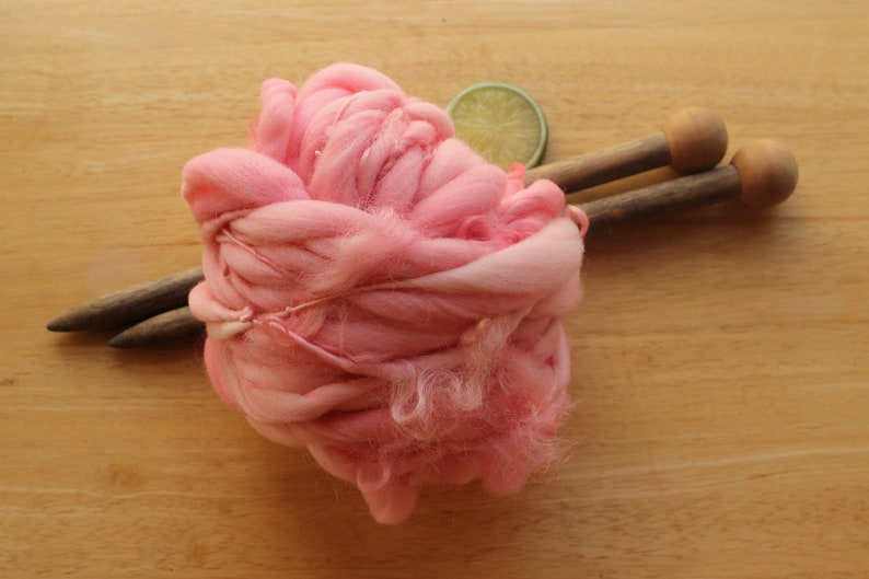 A skein of super bulky, hand dyed, handspun, thick and thin yarn. The yarn is bubblegum pink with wool curls. It is on a light wood background with a pair of large, wooden knitting needles and a lime slice.