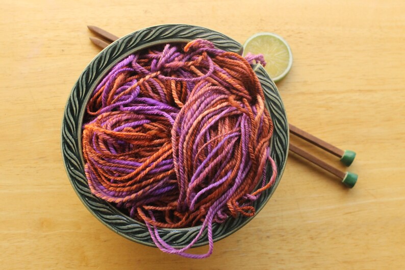 A hank of worsted weight, 2 ply yarn in lavender, peach, and rust. The yarn is nestled in a dark green, ceramic yarn bowl on a light wood background with a pair of square, wooden knitting needles and a lime slice.
