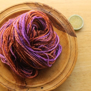 A hank of worsted weight, 2 ply yarn in lavender, peach, and rust. The yarn is piled on a wooden plate on a light wood background with a lime slice.