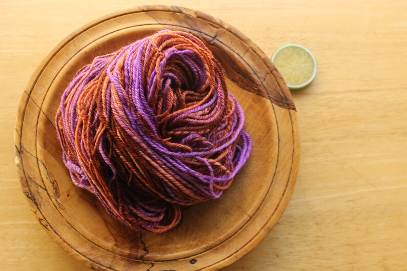 A hank of worsted weight, 2 ply yarn in lavender, peach, and rust. The yarn is piled on a wooden plate on a light wood background with a lime slice.