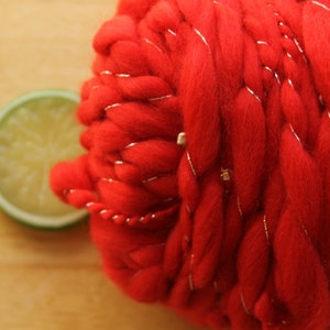 An extreme close up of a skein of handspun, solid red, thick and thin yarn. The super bulky yarn is plied with silver thread and glass beads. It is resting on a light wood background with a lime slice.