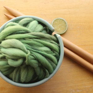 A skein of thick and thin, super bulky, handspun yarn. The yarn is hand dyed in dusty greens. It is nestled in a pale blue, ceramic yarn bowl on a light wood background with a pair of large, wooden knitting needles and a lime slice.