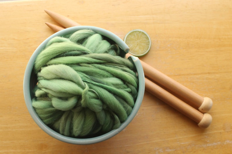 A skein of thick and thin, super bulky, handspun yarn. The yarn is hand dyed in dusty greens. It is nestled in a pale blue, ceramic yarn bowl on a light wood background with a pair of large, wooden knitting needles and a lime slice.