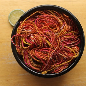 A hank of handspun, worsted weight, 2 ply yarn in red, hot pink, yellow, and white.  The yarn is nestled in a glossy, black, ceramic yarn bowl on a light wood background with a lime slice.
