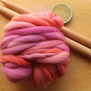A skein of handspun, super bulky, thick and thin yarn. The yarn is hand dyed in self striping lilac and coral. The yarn is resting on a light wood background with a pair of large, wooden knitting needles and a lime slice.