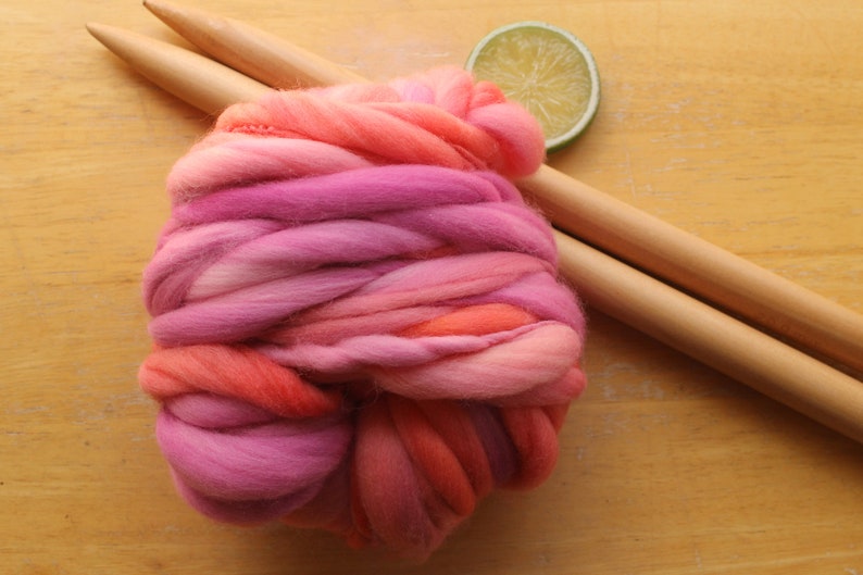 A skein of handspun, super bulky, thick and thin yarn. The yarn is hand dyed in self striping lilac and coral. The yarn is resting on a light wood background with a pair of large, wooden knitting needles and a lime slice.