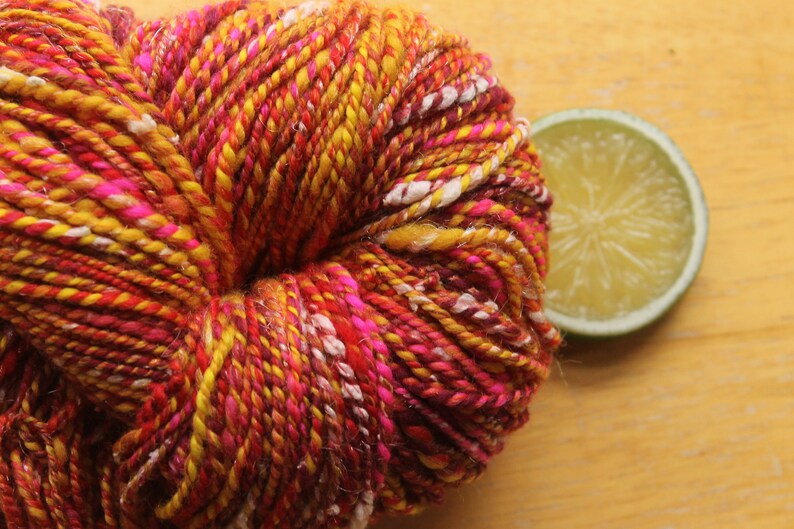 An extreme close up of a skein of handspun, worsted weight, 2 ply yarn in red, hot pink, yellow, and white.  The yarn is resting on a light wood background with a lime slice.