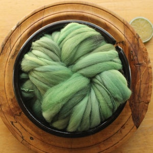 A skein of thick and thin, super bulky, handspun yarn. The yarn is hand dyed in dusty greens. It is nestled in a glossy, black, ceramic yarn bowl, layered on a handmade, wooden plate on a light wood background with a lime slice.