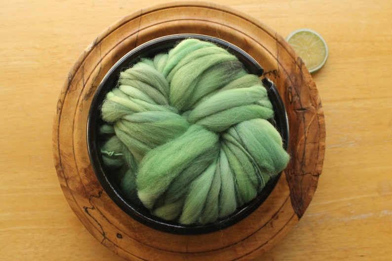 A skein of thick and thin, super bulky, handspun yarn. The yarn is hand dyed in dusty greens. It is nestled in a glossy, black, ceramic yarn bowl, layered on a handmade, wooden plate on a light wood background with a lime slice.