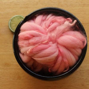 A skein of super bulky, hand dyed, handspun, thick and thin yarn. The yarn is bubblegum pink with wool curls. It is nestled in a glossy, black, ceramic yarn bowl on a light wood background with a lime slice.