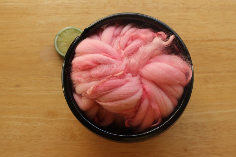 A skein of super bulky, hand dyed, handspun, thick and thin yarn. The yarn is bubblegum pink with wool curls. It is nestled in a glossy, black, ceramic yarn bowl on a light wood background with a lime slice.