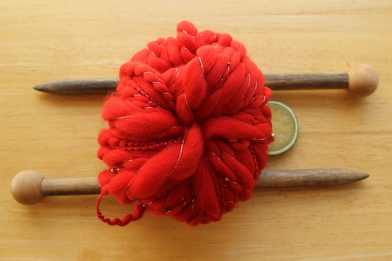 A skein of handspun, solid red, thick and thin yarn. The super bulky yarn is plied with silver thread and glass beads. It is resting on a light wood background with a pair of large, wooden knitting needles and a lime slice.