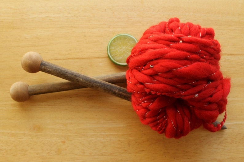 A skein of handspun, solid red, thick and thin yarn. The super bulky yarn is plied with silver thread and glass beads. It is resting on a light wood background with a pair of large, wooden knitting needles and a lime slice.