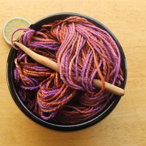 A hank of worsted weight, 2 ply yarn in lavender, peach, and rust. The yarn is nestled in a black, ceramic yarn bowl on a light wood background with a rustic, wooden crochet hook and a lime slice.