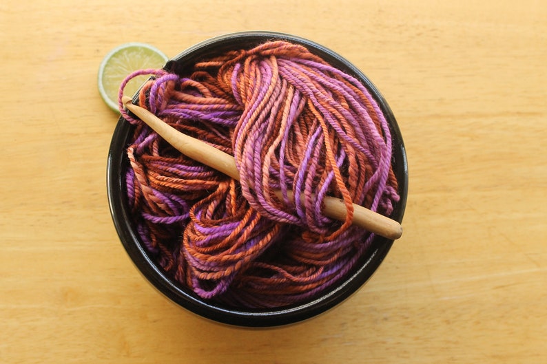 A hank of worsted weight, 2 ply yarn in lavender, peach, and rust. The yarn is nestled in a black, ceramic yarn bowl on a light wood background with a rustic, wooden crochet hook and a lime slice.