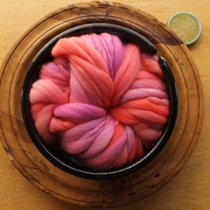 A skein of handspun, super bulky, thick and thin yarn. The yarn is hand dyed in self striping lilac and coral. The yarn is nestled in a ceramic yarn bowl, layered on a wooden plate, on a light wood background with a lime slice.