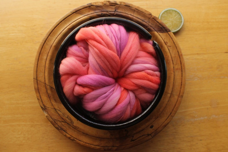 A skein of handspun, super bulky, thick and thin yarn. The yarn is hand dyed in self striping lilac and coral. The yarn is nestled in a ceramic yarn bowl, layered on a wooden plate, on a light wood background with a lime slice.