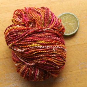 A skein of handspun, worsted weight, 2 ply yarn in red, hot pink, yellow, and white.  The yarn is resting on a light wood background with a lime slice.