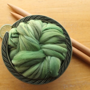 A skein of thick and thin, super bulky, handspun yarn. The yarn is hand dyed in dusty greens. It is nestled in a deep green, ceramic yarn bowl on a light wood background with a pair of large, wooden knitting needles and a lime slice.