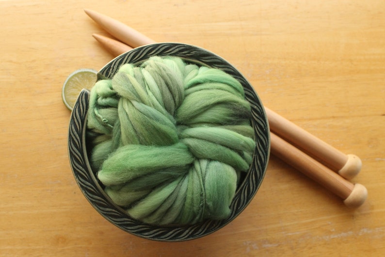 A skein of thick and thin, super bulky, handspun yarn. The yarn is hand dyed in dusty greens. It is nestled in a deep green, ceramic yarn bowl on a light wood background with a pair of large, wooden knitting needles and a lime slice.