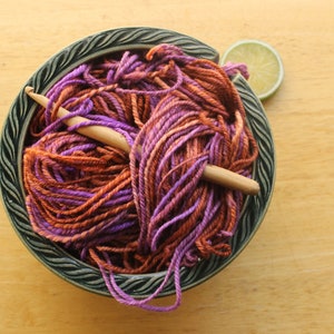 A hank of worsted weight, 2 ply yarn in lavender, peach, and rust. The yarn is nestled in a dark green, ceramic yarn bowl on a light wood background with a rustic, wooden crochet hook and a lime slice.