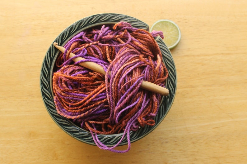 A hank of worsted weight, 2 ply yarn in lavender, peach, and rust. The yarn is nestled in a dark green, ceramic yarn bowl on a light wood background with a rustic, wooden crochet hook and a lime slice.