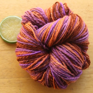 A skein of worsted weight, 2 ply yarn in lavender, peach, and rust. The yarn is on a light wood background with a lime slice.