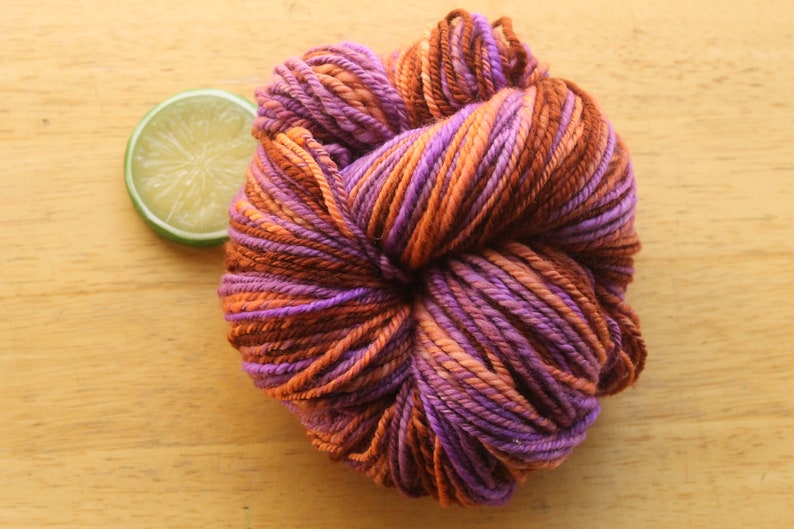 A skein of worsted weight, 2 ply yarn in lavender, peach, and rust. The yarn is on a light wood background with a lime slice.