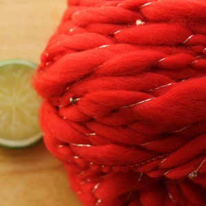 An extreme close up of a skein of handspun, solid red, thick and thin yarn. The super bulky yarn is plied with silver thread and glass beads. It is resting on a light wood background with a lime slice.