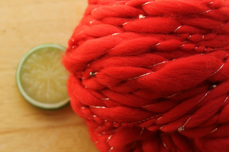 An extreme close up of a skein of handspun, solid red, thick and thin yarn. The super bulky yarn is plied with silver thread and glass beads. It is resting on a light wood background with a lime slice.