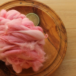 A skein of super bulky, hand dyed, handspun, thick and thin yarn. The yarn is bubblegum pink with wool curls. It is resting on a handmade, wooden plate on a light wood background with a lime slice.