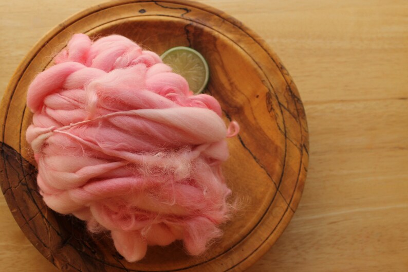 A skein of super bulky, hand dyed, handspun, thick and thin yarn. The yarn is bubblegum pink with wool curls. It is resting on a handmade, wooden plate on a light wood background with a lime slice.