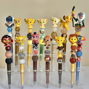 Könnte beinhalten: Ein Set mit 14 bunten Stiften, die mit Perlen und Miniaturfiguren aus dem König der Löwen verziert sind. Die Stifte zeigen Figuren wie Simba, Nala, Timon, Pumbaa und Scar.